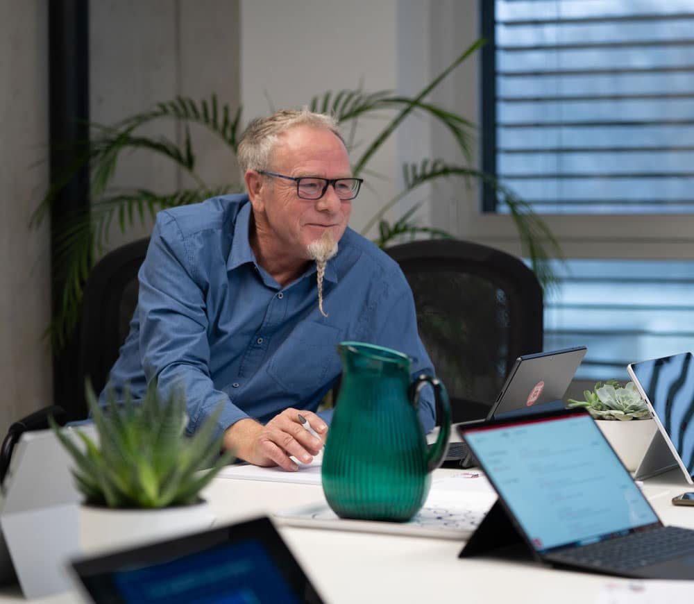 Ein Mann am Tisch mit Laptops und Krug – auf dem Bildschirm: Terminbuchung.